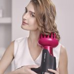Woman styling her hair with pink Panasonic hairdryer attachment for creating curls and volume, wearing white top