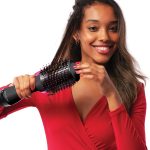 Woman styling hair with Revlon hot air brush, wearing a red top and smiling, showcasing hair care and styling tools