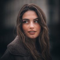 Young woman with long brown hair in a coat looking to the side against a blurred city background
